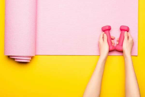 Top view of female hands with dumbbells on pink fitness mat on yellow background — Stock Photo