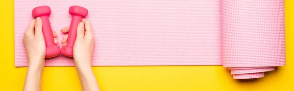 Vue de dessus des mains féminines avec haltères sur tapis de fitness rose sur fond jaune, vue panoramique — Photo de stock