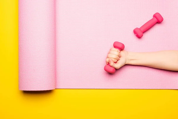 Vista superior da mão feminina com haltere no tapete de fitness rosa no fundo amarelo — Fotografia de Stock