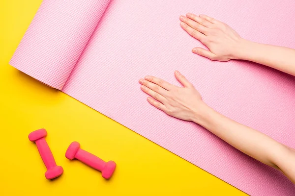 Vista superior de las manos femeninas en la alfombra de fitness rosa cerca de pesas sobre fondo amarillo - foto de stock