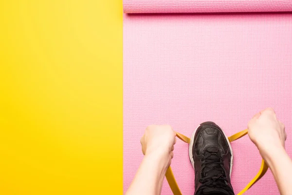 Vista recortada de la mujer de pie en la banda elástica en la alfombra de fitness rosa sobre fondo amarillo - foto de stock