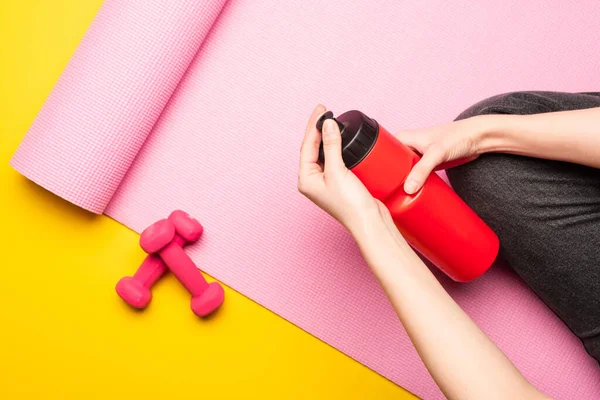 Partial view of woman holding sports bottle while sitting on pink fitness mat near dumbbells on yellow background — Stock Photo