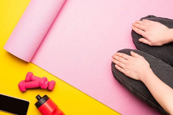Vue partielle de la femme assise sur un tapis de fitness rose près d'une bouteille de sport, smartphone, haltères sur fond jaune — Photo de stock