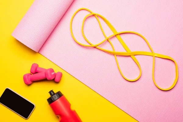 Top view of resistance band on pink fitness mat near smartphone, sports bottle, dumbbells on yellow background — Stock Photo