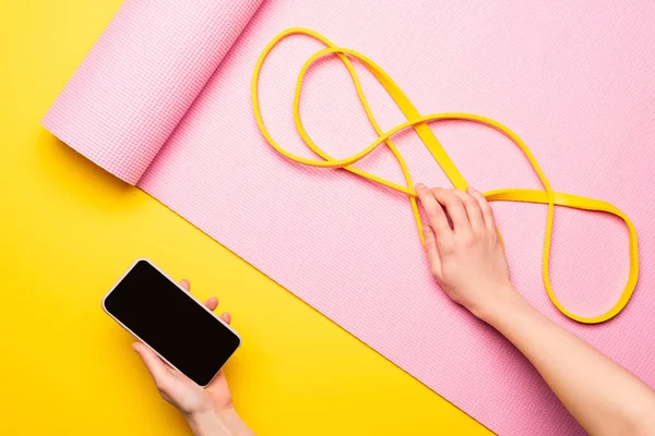 Vue recadrée de la femme tenant smartphone et bande de résistance sur tapis de fitness rose sur fond jaune — Photo de stock