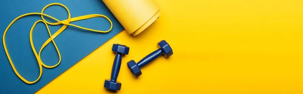 Top view of blue fitness mat with dumbbells and resistance band on yellow background, panoramic shot — Stock Photo