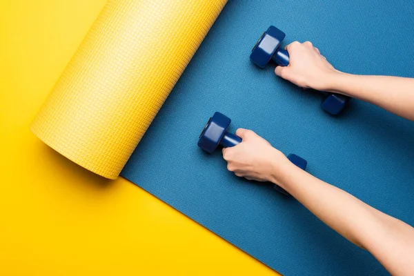 Vista cortada de mulher segurando halteres no tapete de fitness azul no fundo amarelo — Fotografia de Stock