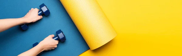 Cropped view of woman holding dumbbells on blue fitness mat on yellow background, panoramic shot — Stock Photo