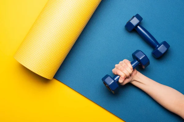 Vista ritagliata della donna che tiene i manubri sul tappeto fitness blu su sfondo giallo — Stock Photo