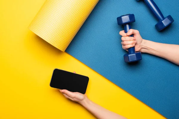 Vista recortada de la mujer sosteniendo la mancuerna y el teléfono inteligente en la estera de fitness azul sobre fondo amarillo - foto de stock