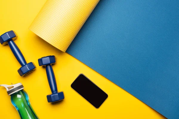 Vista superior de la alfombra de fitness azul con mancuernas, teléfono inteligente y botella de deportes sobre fondo amarillo - foto de stock