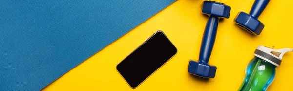 Vista superior do tapete de fitness azul com halteres, smartphone e garrafa de esportes no fundo amarelo, tiro panorâmico — Fotografia de Stock