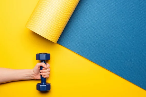 Vista cortada da mulher segurando haltere no tapete de fitness azul no fundo amarelo — Fotografia de Stock