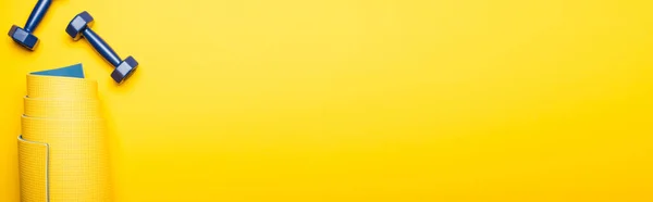 Vista superior de estera de fitness enrollada y pesas azules sobre fondo amarillo, plano panorámico - foto de stock
