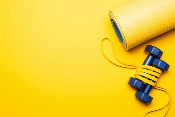 Top view of fitness mat with blue dumbbells and resistance band on yellow background — Stock Photo