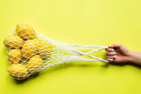 Cropped view of woman holding ripe whole lemons in string bag on yellow background — Stock Photo