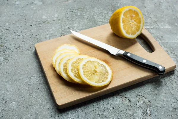 Ripe yellow sliced lemon on wooden cutting board with knife on concrete textured surface — Stock Photo