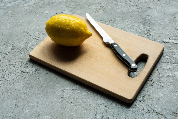 Ripe yellow whole lemon on wooden cutting board with knife on concrete textured surface — Stock Photo