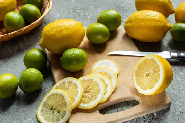 Ripe yellow lemons and limes on wooden cutting board with knife on concrete textured surface — Stock Photo