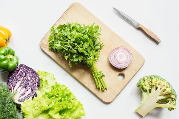 Vue du dessus des légumes mûrs près de la planche à découper en bois avec persil et oignon sur surface blanche — Photo de stock