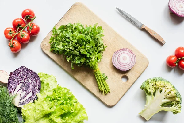 Top view of ripe vegetables and knife near wooden cutting board with parsley and onion on white surface, panoramic shot — Stock Photo