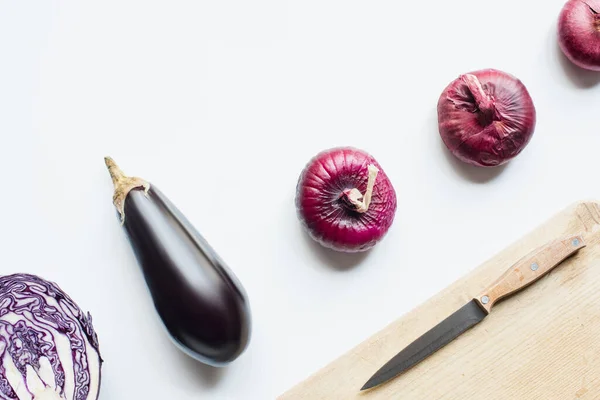 Flat lay with red onion, red cabbage, bargplant near wooden cutting board with knife on white background — стоковое фото