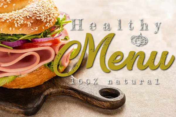 Delicious bagel with ham on wooden cutting board near healthy menu lettering on textured surface — Stock Photo