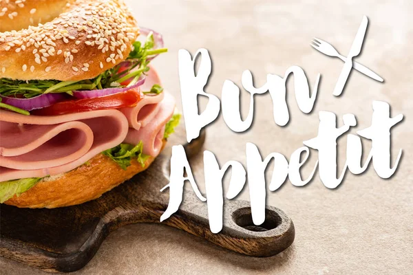 Delizioso bagel con prosciutto su tagliere di legno vicino bon lettering appetit su superficie testurizzata — Foto stock