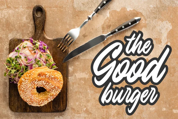 Top view of fresh bagel with meat on cutting board with cutlery near the good burger lettering on textured surface — Stock Photo