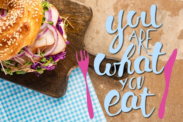 Top view of fresh bagel with meat on cutting board and plaid napkin near you are what you eat lettering and cutlery illustration — Stock Photo