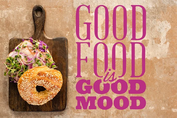 Top view of bagel with meat on cutting board with cutlery near good food is good mood lettering on textured surface — Stock Photo