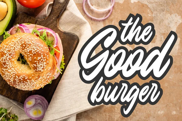 Vista superior de bagel na placa de corte de madeira e guardanapo de pano perto do bom hambúrguer lettering no fundo texturizado — Fotografia de Stock