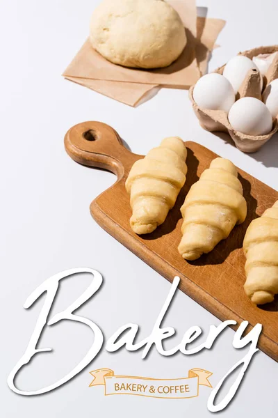Raw croissants on wooden cutting board near eggs, dough, bakery and coffee lettering on white — Stock Photo