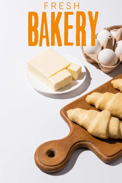 Raw croissants on wooden cutting board near butter, eggs and fresh bakery lettering on white background — Stock Photo