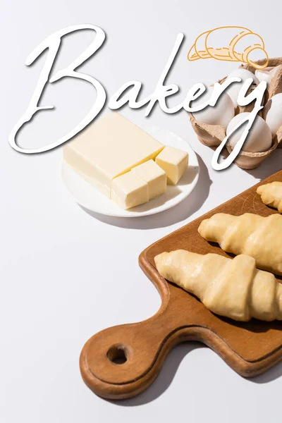 Croissants crus sur planche à découper en bois près du beurre, oeufs et lettrage boulangerie sur fond blanc — Photo de stock