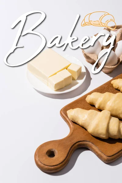 Raw croissants on wooden cutting board near butter, eggs and bakery lettering on white background — Stock Photo