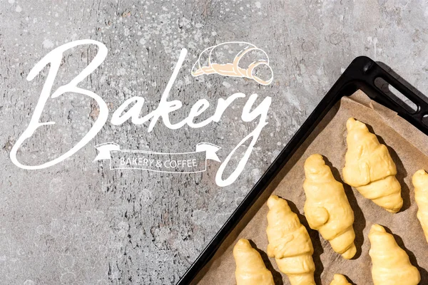 Top view of uncooked croissants on baking tray near bakery and coffee lettering on concrete grey surface — Stock Photo