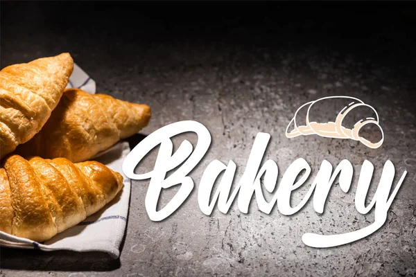 Fresh baked croissants on towel near bakery lettering and illustration on concrete grey surface — Stock Photo