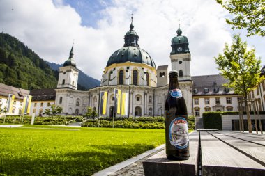A bottle of Ettaler beer in front of the main facade of of Ettal Abbey (Kloster Ettal), a Benedictine monastery in the village of Ettal, Bavaria, Germany clipart