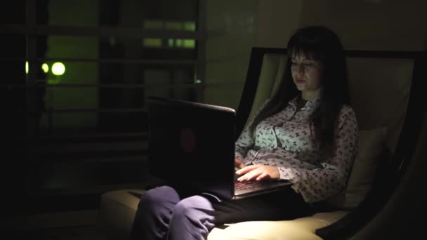 Chica sentada junto a la ventana con un ordenador portátil en la noche — Vídeos de Stock