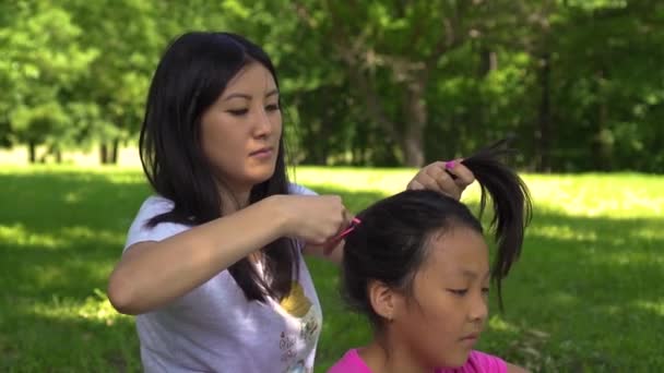 Young asian mom does her daughters hair in the park, day — стоковое видео