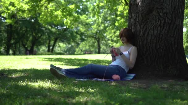 Ung flicka gör stickning i parken under ett träd — Stockvideo