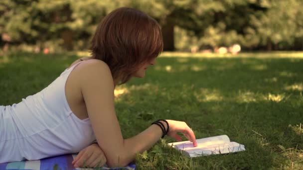 Menina deitada na grama no parque e lendo um livro — Vídeo de Stock