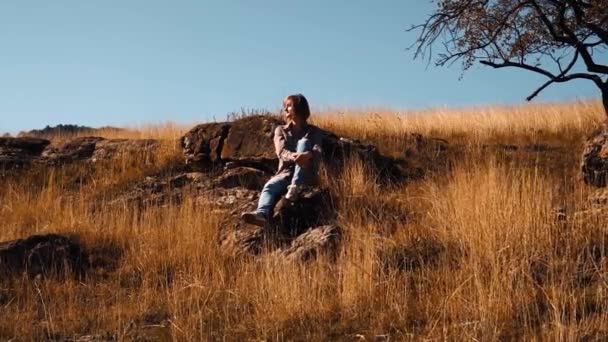 Chica se sienta en otoño en una piedra cerca de un árbol — Vídeos de Stock