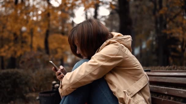 Молодая девушка сидит одна в парке с телефоном — стоковое видео