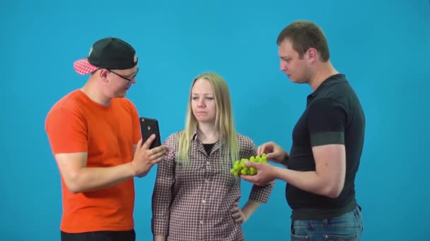 Dwóch facetów zabawia jedną dziewczynę winogronami i smartfonem — Wideo stockowe