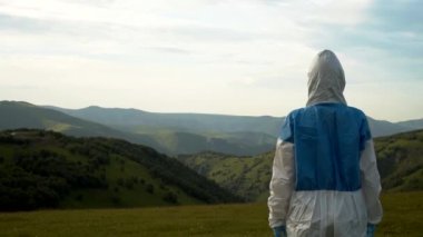 Koruyucu biyolojik kıyafetli bir adam dağlarda dikiliyor.