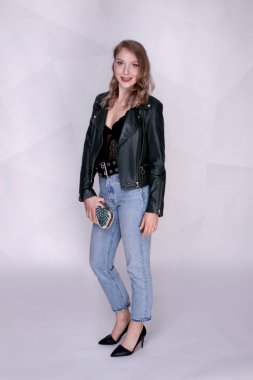 Güzel bir kız beyaz bir arka plan üzerinde duruyor, o kot pantolon ve deri ceket ve siyah bir vücut giymiş. Elinde küçük bir çanta tutuyor. Kız sarışın..