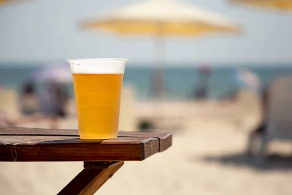 Wet glass of golden cool tasty beer on the wooden table on the shore of the sea or ocean on the sunset. Concept of beach bar or party. Alcohol beverage.