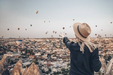 Şapkalı genç, arka planda uçan hava balonları ile dağda duruyor. Gün doğumunda kızı işaret eden parmak. Arkadan bak. Ünlü turistik Türk bölgesi Kapadokya.